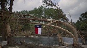 A Texas flag hangs from a flood-damaged tree on the bank of the Guadalupe River in Kerrville, Texas after deadly floods in July 2025. A group of emergency experts appointed by President Trump is recommending that the Federal Emergency Management Agency (FEMA) provide less money to states to help prepare for and respond to disasters including floods.