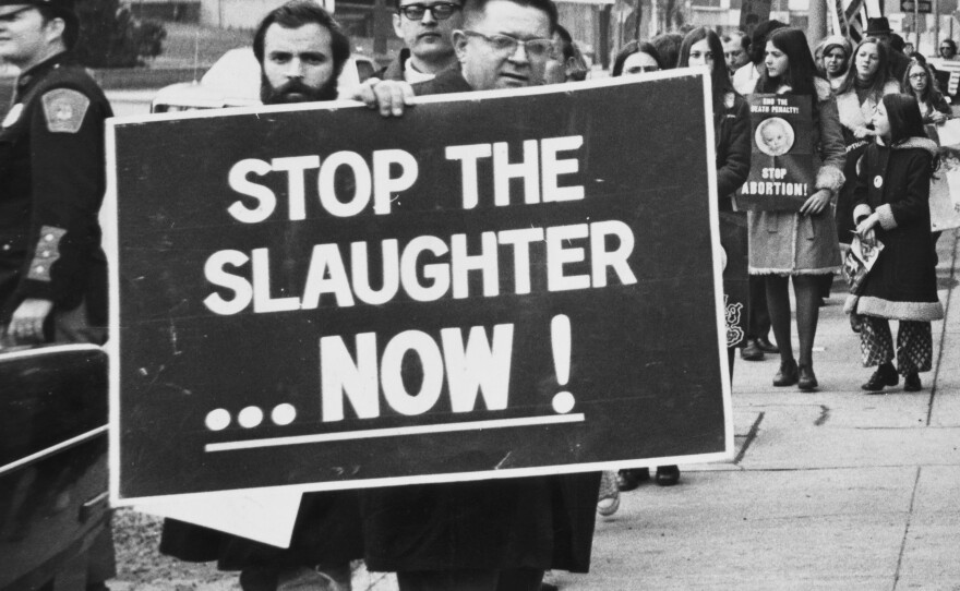 Members of a Right to Life committee holding a banner reading 'Stop the slaughter now!' and a placard reading 'The Supreme Court Injustice' during a protest, location unspecified, 1974.