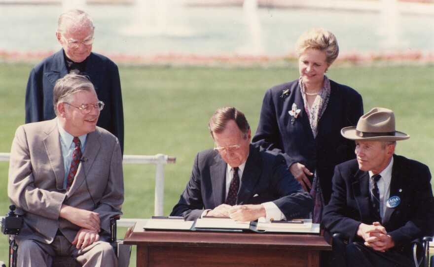 President George H. W. Bush (center) signs the Americans with the Disabilities Act during a ceremony in the White House Rose Garden on July 26, 1990 with (seated, from left to right) Evan Kemp, Chairman of the Equal Employment Opportunity Commission, and Justin Dart, Vice Chair of the National Council on Disability.