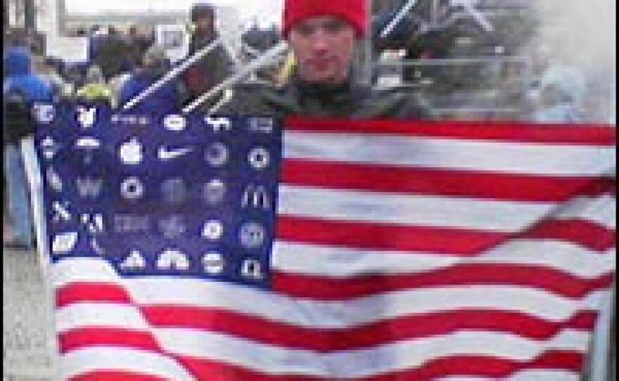 A protester in a designated protest area along Pennsylvania Avenue holds a flag with corporate logos in place of stars.