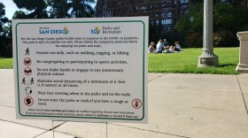 A sign at Balboa Park telling visitors what's allowed at the park while a family is having a picnic in the background on June 14, 2020, the first weekend the park reopened to visitors.