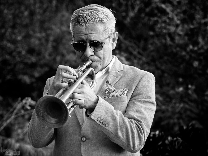 Gilbert Castellanos plays the trumpet in a black and white, undated photo. He is standing outside against a leafy background, and is wearing a light-colored suit jacket and sunglasses.