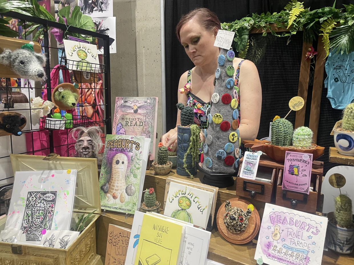 Asia Estigoy is shown at the ToshWerks booth at San Diego Comic-Con in 2024, with a variety of handmade comics, pins, crocheted cacti and taxiderm