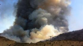 A large plume of white and black smoke rises from a hillside brush fire in San Marcos.