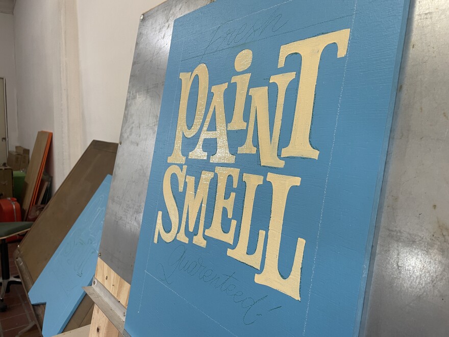 An in-progress sign is shown at Koy Sun's artist residency studio in Bread & Salt on Oct. 4, 2024. It's a blue sign with cream lettering reading PAINT SMELL, with pencil lettering showing Fresh and Guaranteed on the top and bottom.