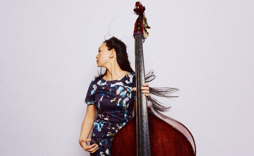 Bassist Linda May Han Oh holds a large upright bass in one hand, and looks off to the side. Her long, brown hair is flipping outward, as if she is spinning.