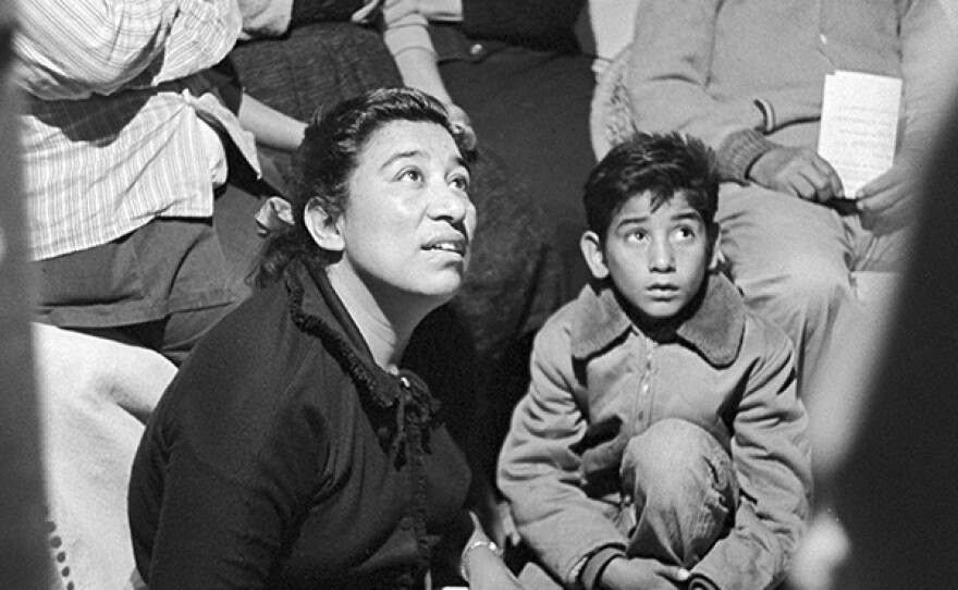 Migrant mother Maria Moreno became the first farmworker woman in America to be hired as a union organizer. Surrounded by farmworkers and their children at a house meeting, Maria listens and takes notes. 1960