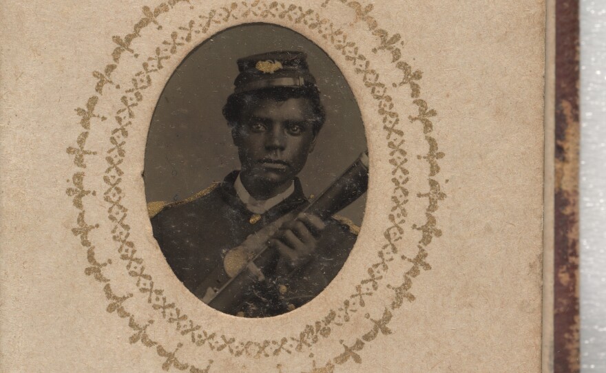 A black-and-white photograph of James Tall. He is wearing a buttoned-up jacket with epaulets on the shoulders and a kepi. He is holding a rifle and his left hand is visible in the picture. He is leaning slightly to the right side of the picture.