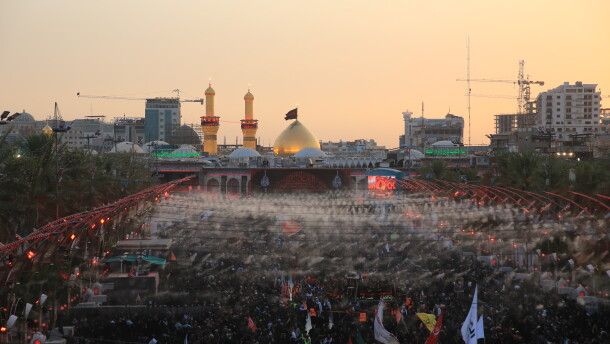 Large crowds of pilgrims fill the area between the two holy shrines in Karbala at sunset, with the illuminated dome and minarets of Imam Hussein's shrine visible in the background during the Arbaeen commemoration.
