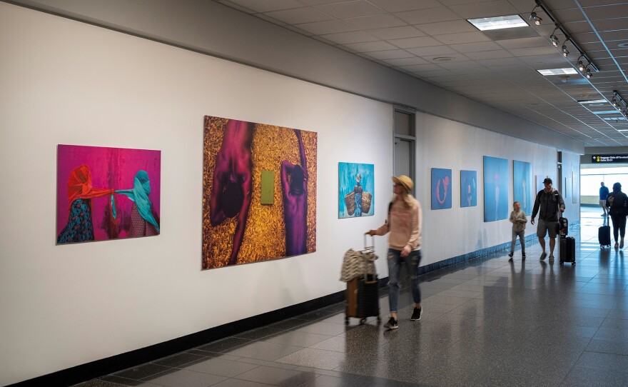 Travelers wheel suitcases past large, brightly colored paintings on the wall. The largest painting features two men lying face-down on a yellow surface, with a green square in front of them. They are not wearing shirts, and the yellow background looks like it could be surreal water. Their brown skin is rendered a deep purple in the ultra-saturated colors. 