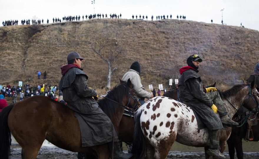 Protesters gather at Standing Rock Reservation on Thanksgiving Day to build a bridge to Turtle Island, which they consider sacred ground. Police are seen lining the island hill beyond them.