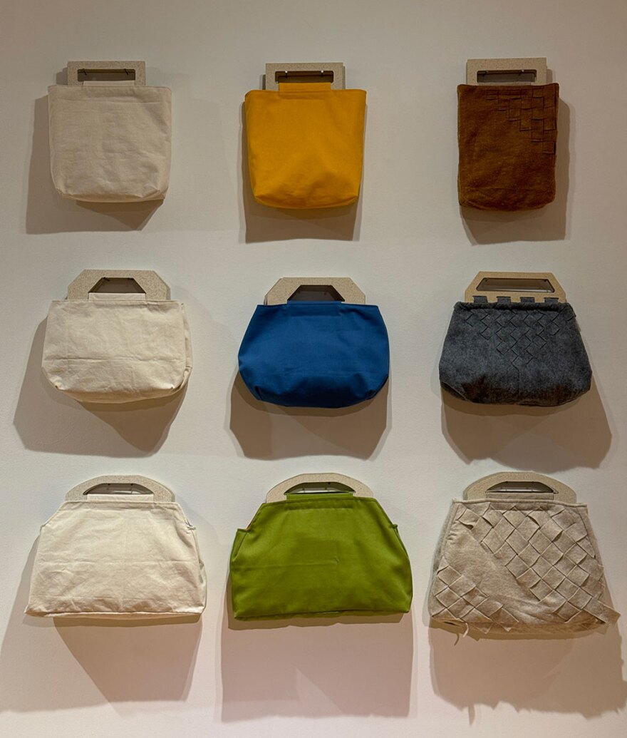 Medium, artist-made bags are shown attached to the wall in three rows of three. The bags are made with a variety of colors and textured materials, but all have a wooden-composite handle.