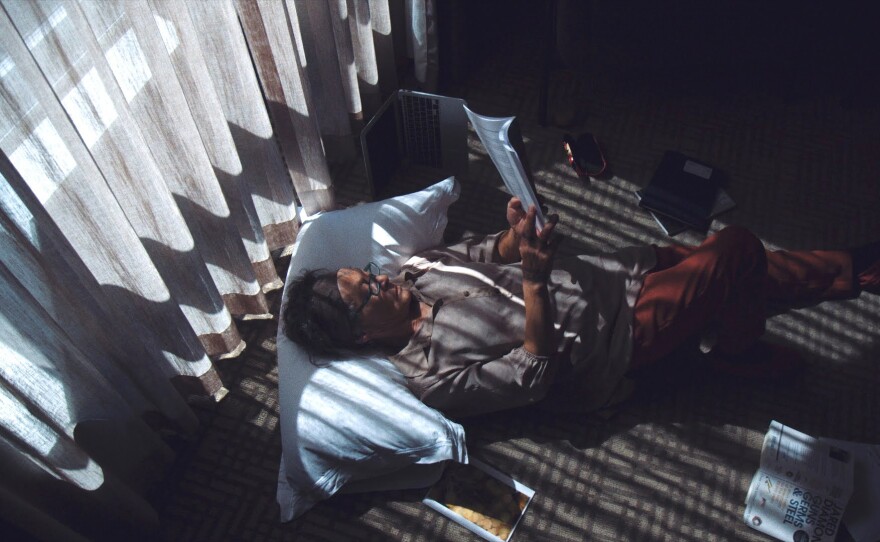 Aaju Peter lies on a brown carpet in front of a bright window with a long gauzy grey curtain in front of it.