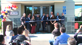 A group of men and women stand in front of STEP's new headquarters holding a long blue ribbon while two in the middle cut it with scissors.