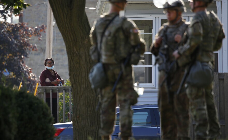 A woman looks on from a home as the Wisconsin National Guard protect a municipal building in Kenosha.