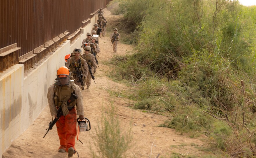 Marines with 1st Combat Engineer Battalion, armed with rifles and chainsaws, some dressed in blaze orange helmets and pants, walk in a column along the border fence move into position along the southern border to conduct vegetation clearance operations near Yuma, Ariz., Dec. 18, 2025.