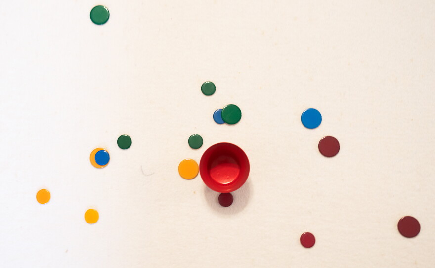 A tiddlywinks game mid-play, with winks spread out around the pot. Though players eventually want to "pot" their "winks," players also strategize how to block their opponents by landing their piece on top of another's piece.