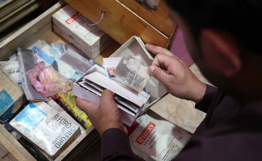 A photo studio owner rummages through boxes of uncollected or duplicate photographs in a dusty backroom.