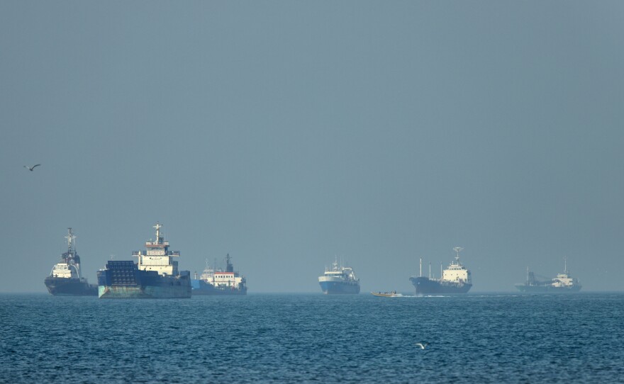 Cargo ships, including bulk carriers and general cargo vessels, sit at anchor offshore as a small motorboat passes in the foreground, in the Strait of Hormuz off Bandar Abbas, Iran, Monday, May 4 , 2026.