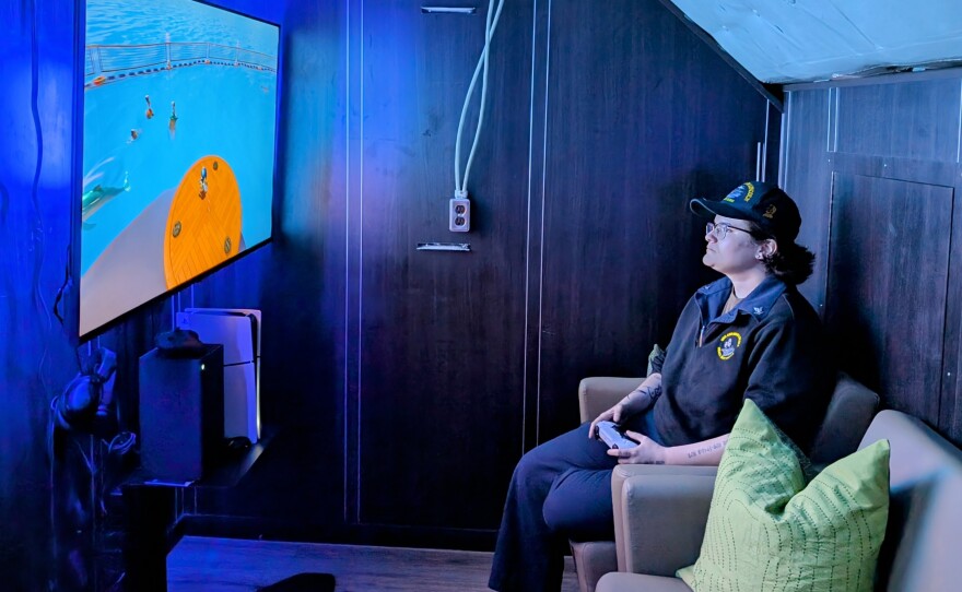 Operations Specialist 3rd Class Dyaal Smith sits in a darkened room illuminated by the light of a large LCD TV. She's playing a game on one of several Playstation 5s available at the USO Center on board the USS Abraham Lincoln on July 8, 2024.