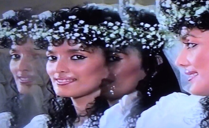 A young Rozana dressed in a white wedding gown looks into the camera through a mirror. There is an effect on the lens which splits the reflection into three. Rozana wears heavy Kohl eyeliner and she has a faint smile at the edge of her lips.