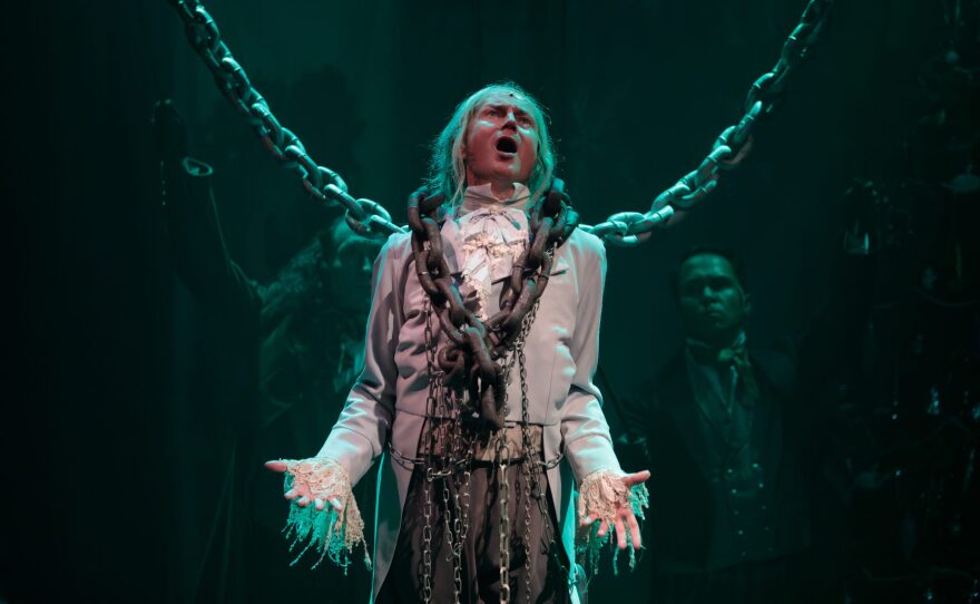 An actor is dressed as the ghost of Jacob Marley in a stage play production of A Christmas Carol. He's wearing white and gray, with frilly sleeves, and is bound by oversized chains. He has long gray hair and a pained expression on his face.