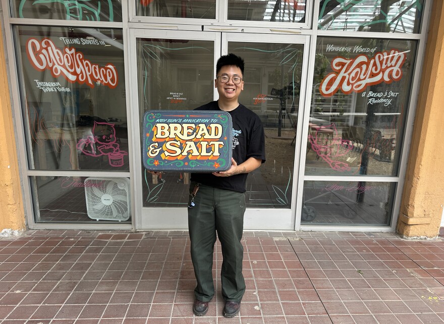 A man holds an old suitcase that's painted like a retro sign, with the words Bread & Salt on the front in vivid colors.