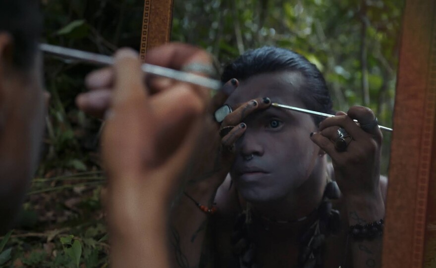 A reflection of Uýra's face is visible as they apply makeup before a mirror. The mirror's frame is golden brown wood and stands out against the dark green of the forest. In the foreground we see the right side of Uýra's face from the back, the right hand holds a long slender brush. From the reflection, viewers see Uýra brush makeup to the left eyebrow, the left eye is open wide staring intently into the mirror while the right hand obscures part of the face. Their face is dusted with a light coat of white paint, a nosering hangs in the septum and their black nailed fingers sport rings, a beaded bracelet visible on the left wrist.