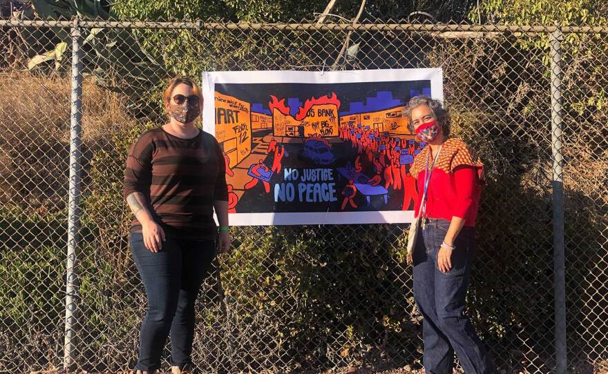 Alessandra Moctezuma and Jenny Armer stand with a work by Michael Chavez, "No Justice No Peace," during the November 10, 2020 installation of Mesa College Art Gallery's forthcoming drive-thru art exhibition.