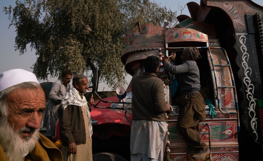 People make adjustments to some of the window elements on a truck. Additional pieces are soldered on to create decorative framing, while other people work on buffing the front of the vehicle and adding ornate stickers to the front bumper.