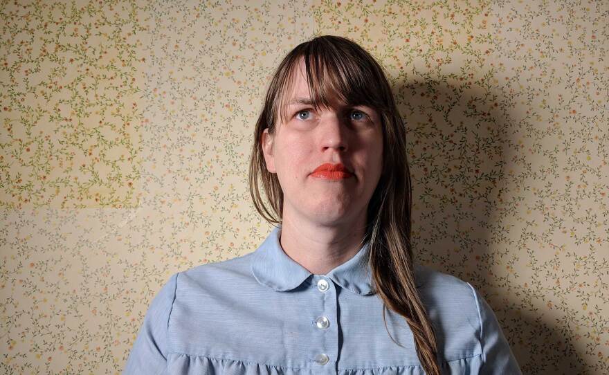 Composer Sarah Hennies is shown against a yellow wallpaper background. She is wearing a blue collared shirt and her long, medium-brown hair is down, falling over one shoulder. She is wearing a coral colored lipstick, and she is not smiling but looking slightly behind the camera.