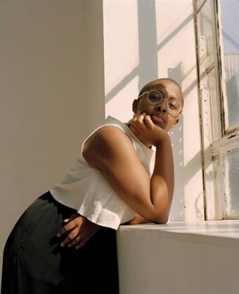 Cécile McLorin Salvant leans against a windowsill, with one hand under her chin. She wears a black skirt, white tank top, and round, light-framed glasses. She has dark skin and her hair is cut close to her head.