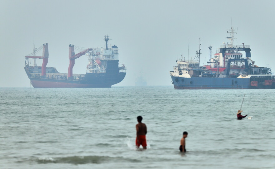 Children wade in the water with cargo ships at anchor in the background and a fisherman nearby, in the Strait of Hormuz off Bandar Abbas, Iran, Tuesday, June 30, 2026.