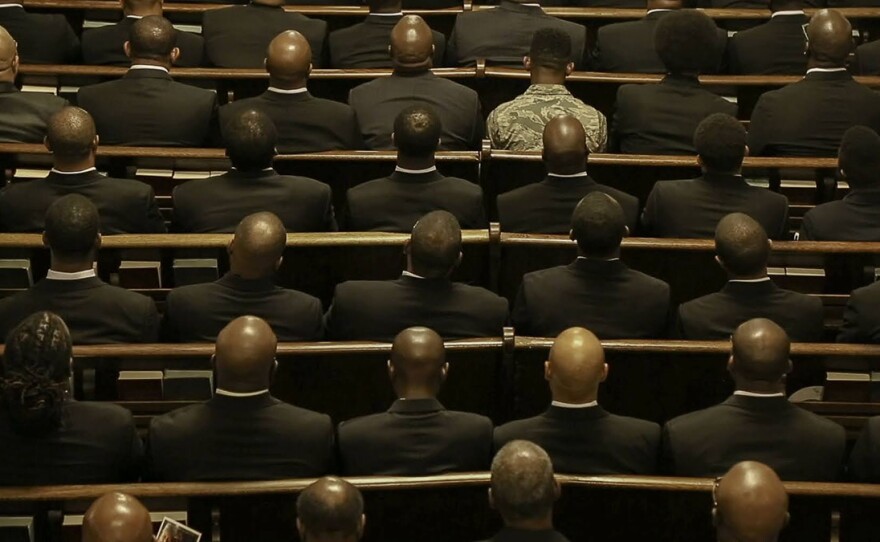 Rows of men in dark suits are seated in church pews, with their backs facing the camera.
