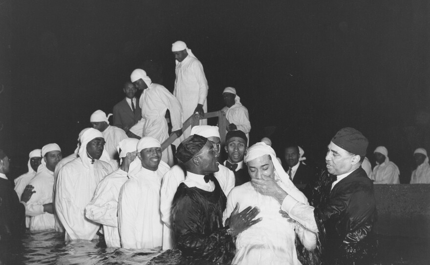 Baptismal candidates in white robes are being led into the water while Elder Michaux and an assistant apparently are raising a candidate immediately after his baptism.