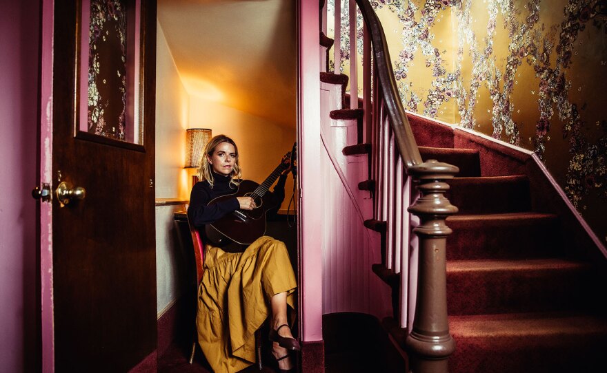 Musician Aoife O'Donovan is shown in an undated photo. She is seated in a small room beneath a winding, wooden staircase, surrounded by pink and yellow walls. She is wearing a black sweater and bright yellow skirt and holding a guitar.