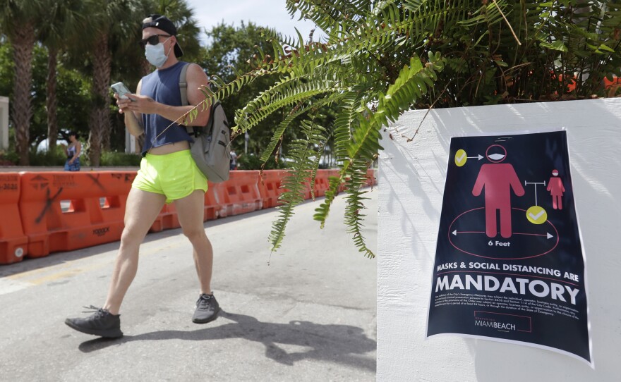 A pedestrian in a mask passes a sign urging people to practice social distancing, on Saturday in Miami Beach, Fla. Just as residents flocked outside to enjoy the Fourth of July, states such as Florida were reporting skyrocketing numbers of confirmed coronavirus cases.