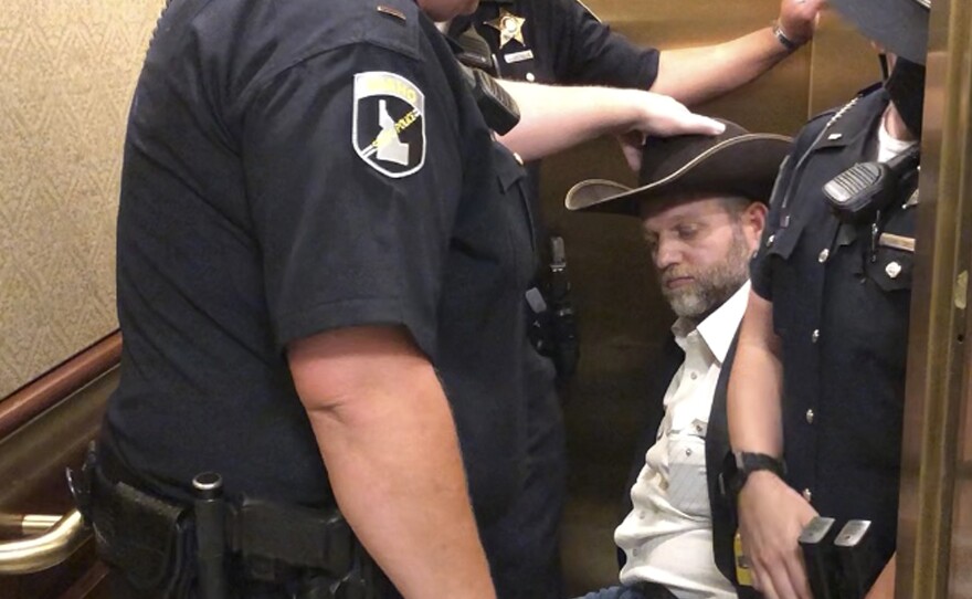 This image taken from video shows anti-government activist Ammon Bundy, center, being wheeled into an elevator in a chair following his arrest at the Idaho Statehouse in Boise, Idaho, Aug. 25.