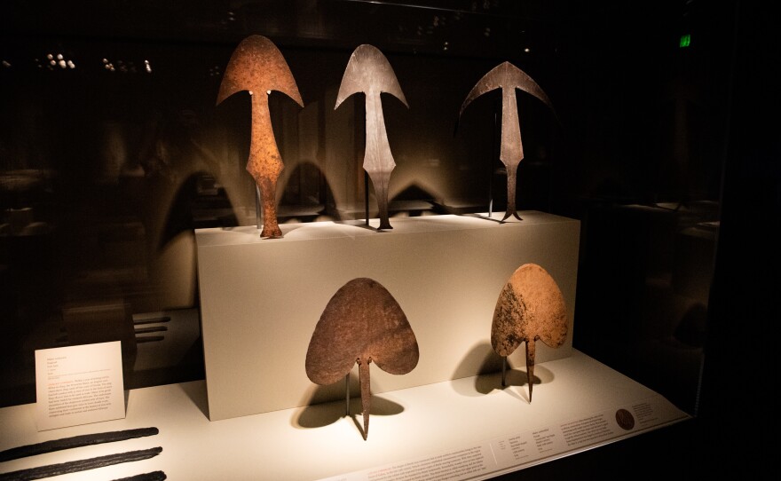Five examples of iron currency from African blacksmiths and European forgers. The forged items are in the middle on the top shelf and at right on the lower level. An African blacksmith made the item on the top shelf at right, introducing more elaborate designs to discourage forgery.