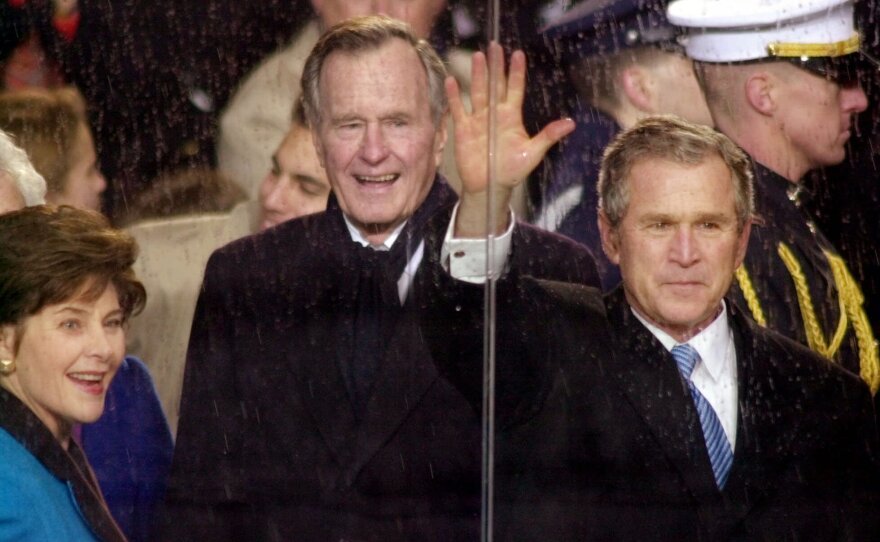 In this Jan. 20, 2001, file photo, standing in the rain, President George W. Bush waves as he watches his inaugural parade pass by the White House viewing stand in Washington, Saturday afternoon, Jan. 20, 2001. With him are his wife and first lady Laura Bush and his father, former President George H.W. Bush.