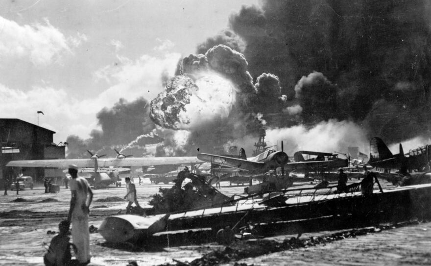 An explosion at the Naval Air Station, Ford Island, Pearl Harbor during the Japanese attack. Sailors stand amid the wreckage, watching as the USS Shaw explodes in the center background. The USS Nevada is also visible in the middle background, with her bow headed toward the left.