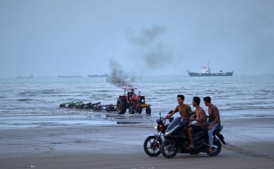 Young men ride motorcycles along the shore as a tractor pulls equipment from the water and commercial vessels appear anchored in the Strait of Hormuz off Bandar Abbas, Iran, Monday, July 27, 2026.