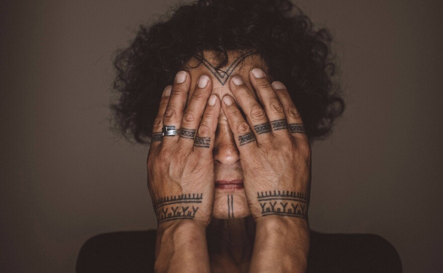 Aaju Peter, an Inuit woman with curly dark hair and a tattooed chin and forehead conceals her eyes behind tattoo-adorned hands against a brown background.