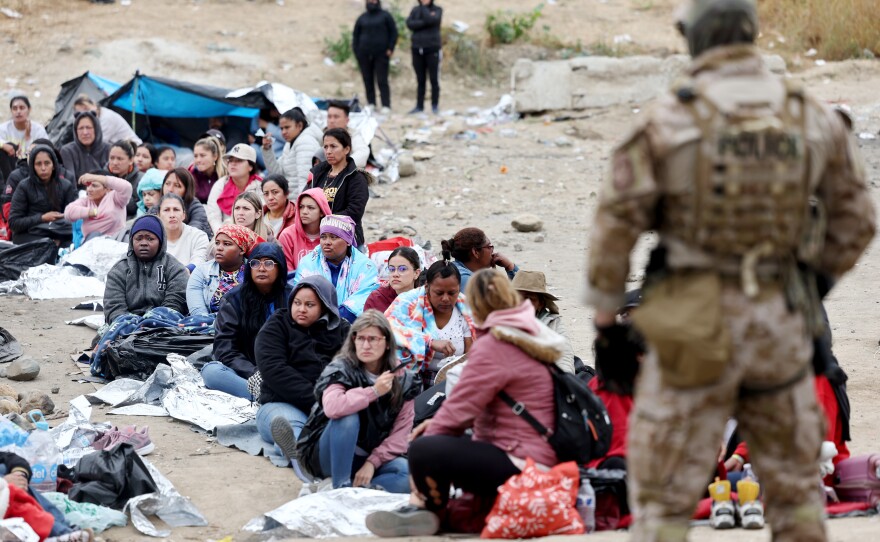 Migrants in a makeshift camp between U.S. and Mexico border walls sit as a Customs and Border Protection officer keeps watch.