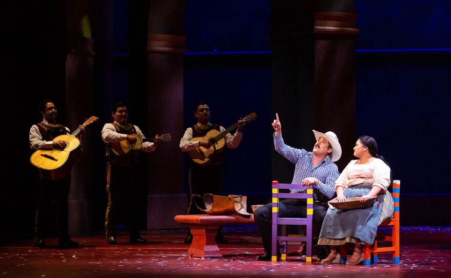 A production of "El Milagro del Recuerdo" shows a man and a woman, seated, pointing at the sky. Three mariachi performers stand in the background, holding guitars.