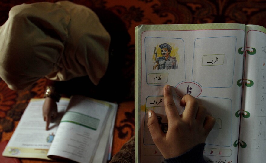 A student reads inside her home in Srinagar, India, as her sister points to a sketch resembling a male police officer in a first-grade textbook
