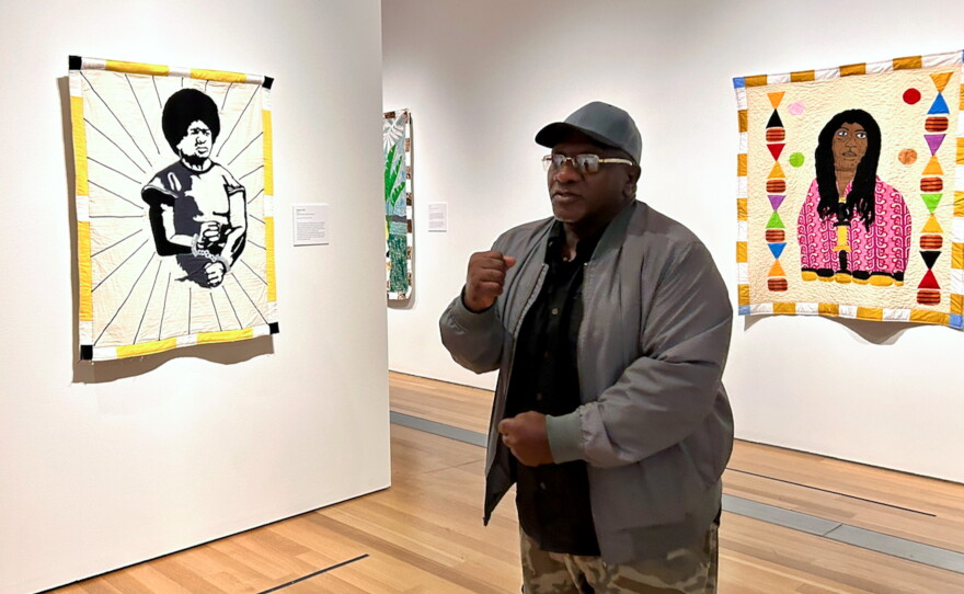 Artist Gary Tyler stands before his quilt, Defiant, 1976, (2023) created in 2023. Tyler says the gesture he is making with his hands was intended to inspire hope and resistance for the people who were protesting the circumstances of his trial.