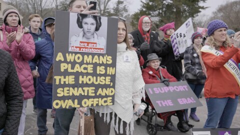 People join together holding signs. "A woman's place in in the house, senate and oval office" and "I support the Era"