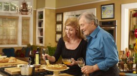 Jacques Pépin enjoys a glass of wine while his daughter, Claudine glazes a tart.