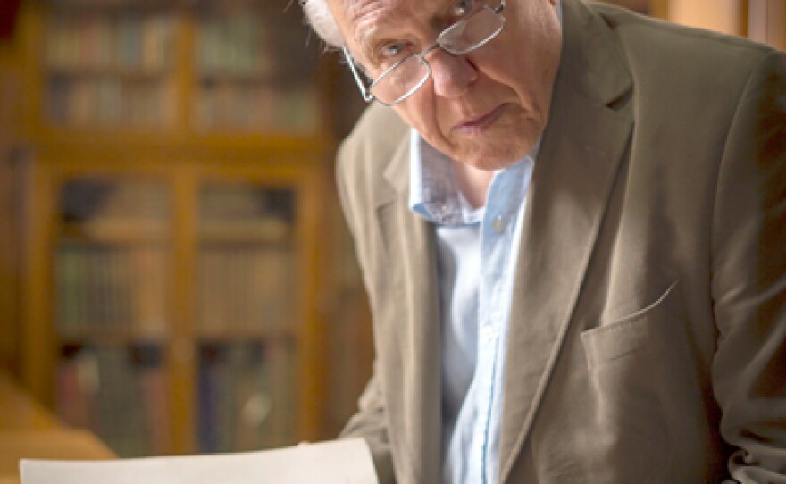 Sir David Attenborough with a book opened to a print of a flightless bird called a cassowary.